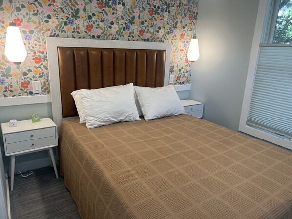 Un lit bien fait, recouvert d’une courtepointe marron, avec deux oreillers blancs et une tête de lit à lattes verticales. On y trouve deux tables de chevet avec des lampes, une horloge et une prise électrique. La pièce est décorée d’un papier peint à motifs floraux et possède une fenêtre avec des stores.