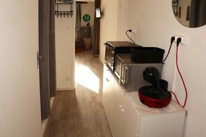 A narrow hallway with a microwave and a red appliance on a counter, leading to a room with a bed and a window.