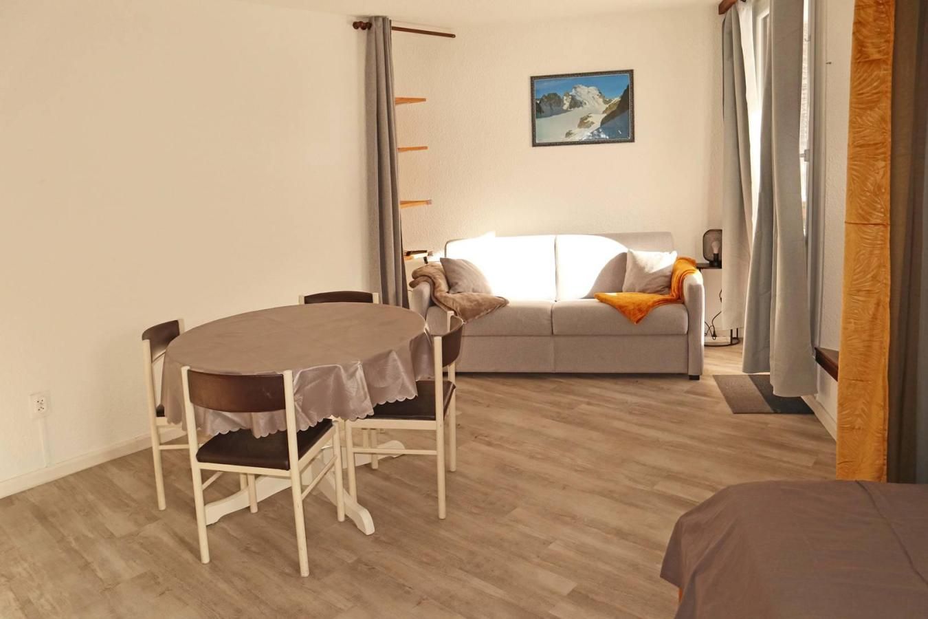 Een kamer met een eettafel en stoelen, een bank, een schilderij aan de muur en een raam met gordijnen.