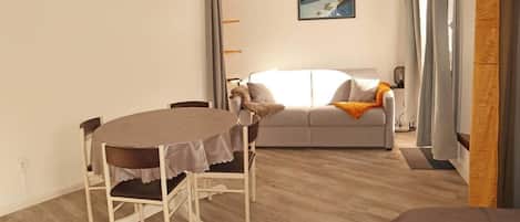 A room with a dining table and chairs, a sofa, a painting on the wall, and a window with curtains.