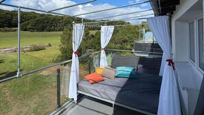 Un balcón con cama, cortinas blancas atadas con lazos rojos, y vista a un paisaje verde.