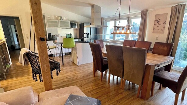 Una sala de estar moderna con sofá, mesa de comedor y sillas, una cocina al fondo y un ventanal con cortinas.