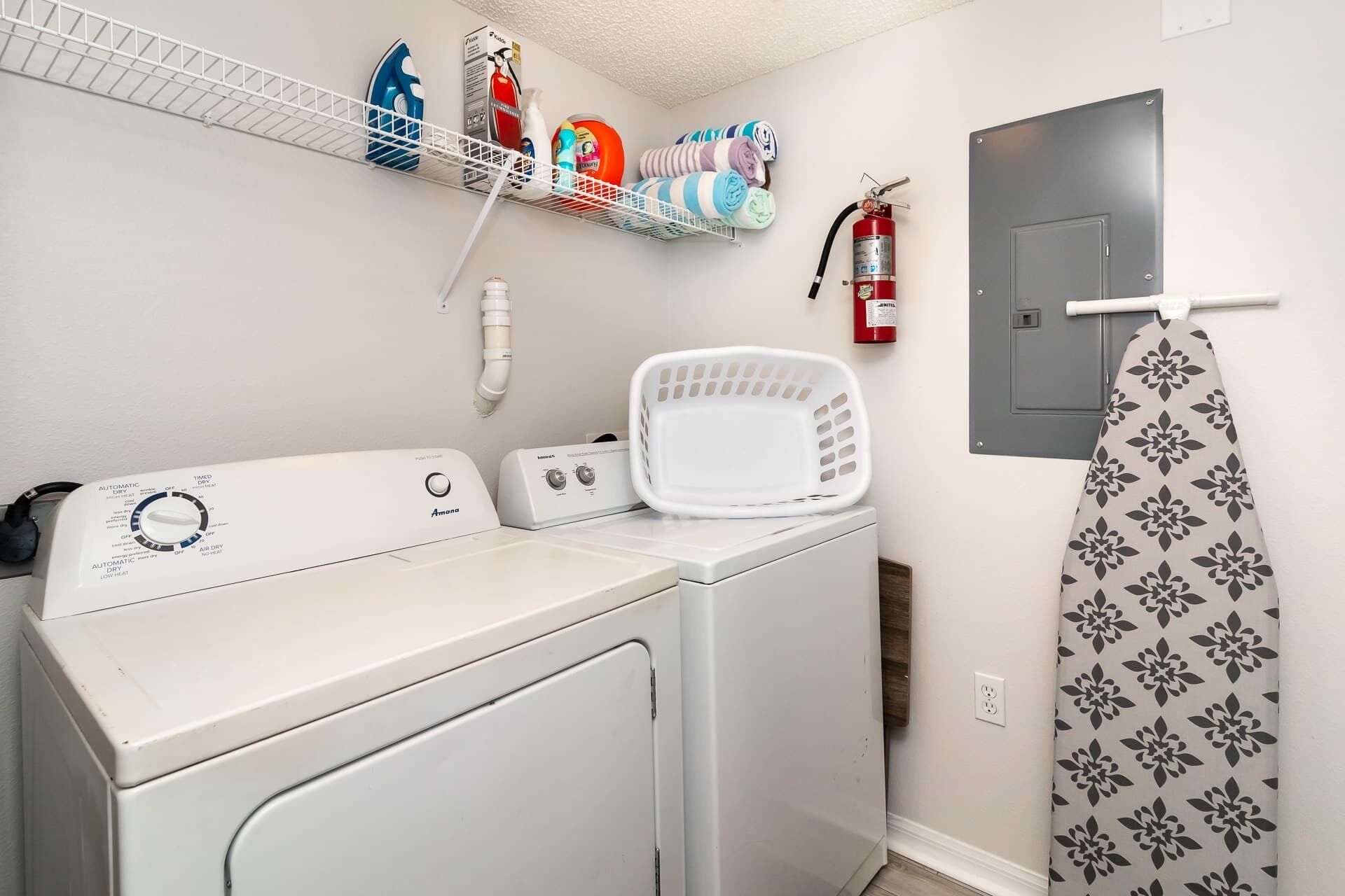 A laundry room with a front-loading washer and dryer, a shelf with cleaning supplies, a fire extinguisher, and a patterned towel hanging on a rack.