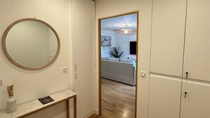 A hallway with a wooden floor, a round mirror, a white console table, and a doorway leading to a living area with a sofa and a hanging light fixture.
