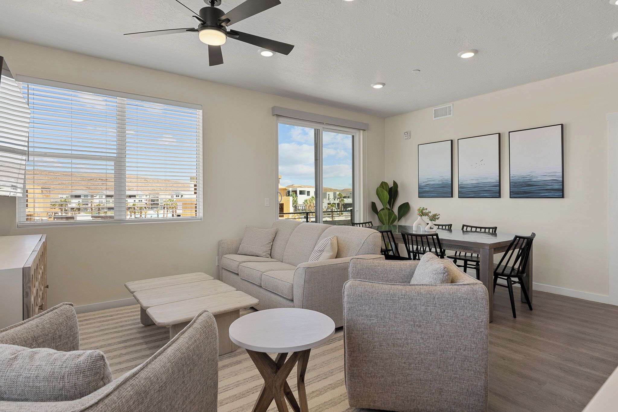 A spacious living area with a sofa, armchairs, a dining table with chairs, and large windows with blinds.