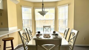 Comedor con una mesa lista para cuatro personas, sillas blancas, lámpara colgante y ventanas amplias con persianas.
