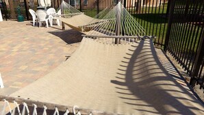 Una hamaca instalada en un patio empedrado, con una valla de metal y sillas de plástico blanco al fondo.