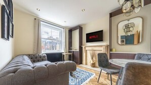 A modern living room with a grey sofa, a dining table with chairs, a fireplace, and a TV mounted on the wall.