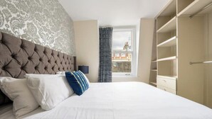 A bedroom with a tufted headboard, a large bed with white bedding, a blue and yellow pillow, a nightstand with a lamp, a window with curtains, and a wardrobe.