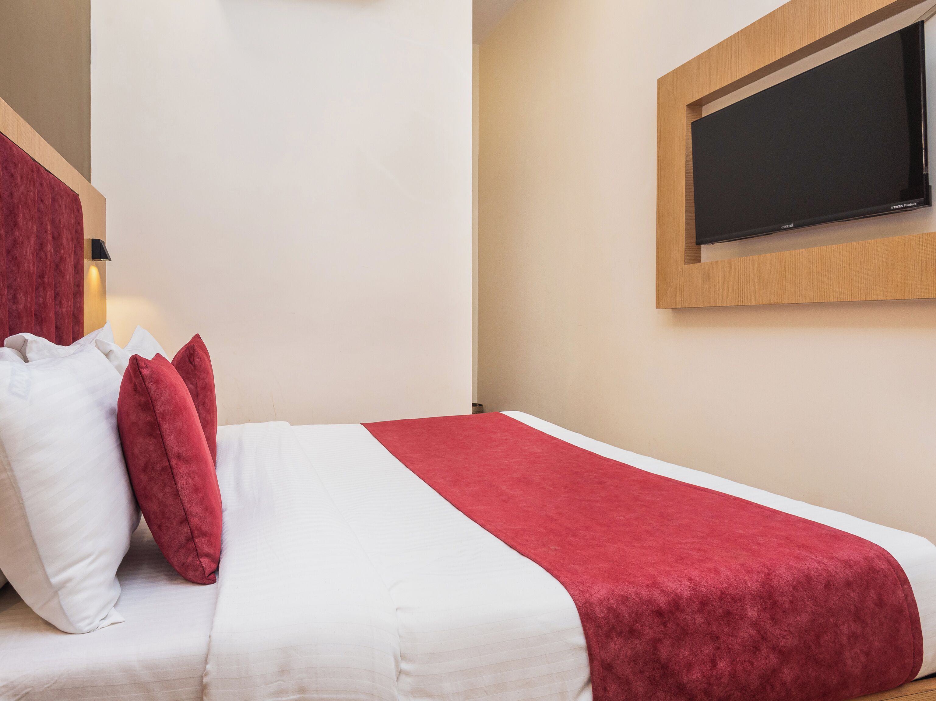 Una cama bien hecha con ropa de cama blanca y roja, cabecera de madera y televisión montada en la pared.