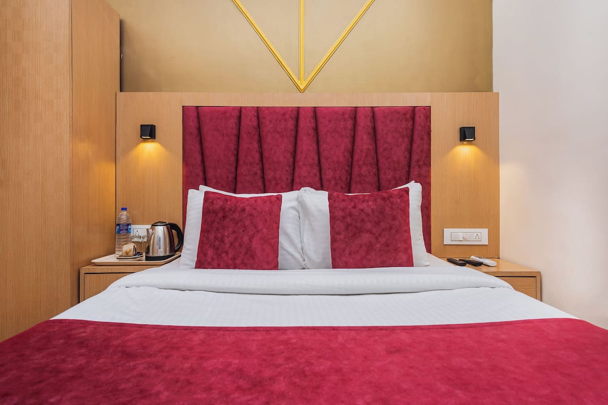 A hotel room with a wooden headboard, a red and white bedspread, a water bottle, a kettle, and a power outlet on the nightstand.