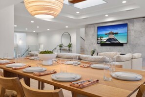 Un espace repas moderne avec une table en bois dressée pour quatre personnes, un grand saladier rempli de verdure et un tableau représentant un paysage tropical accroché au mur.