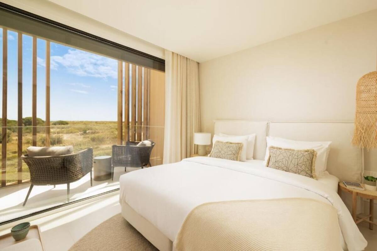 Un dormitorio amplio con una cama grande, vistas al campo a través de ventanales del suelo al techo y un balcón con mobiliario de mimbre.