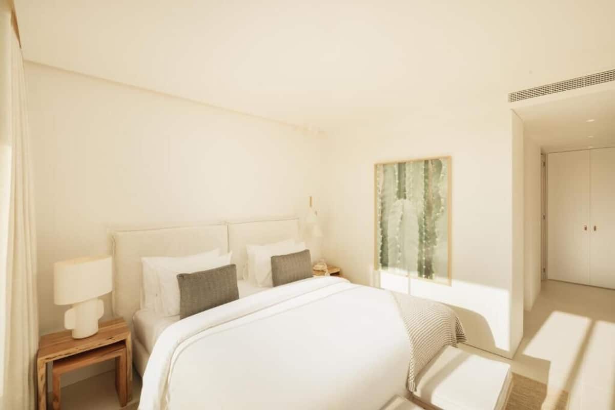 Una habitación de hotel moderna con una cama grande, una mesita de noche con lámpara, una pequeña mesa y un ventanal con vistas a zonas verdes.