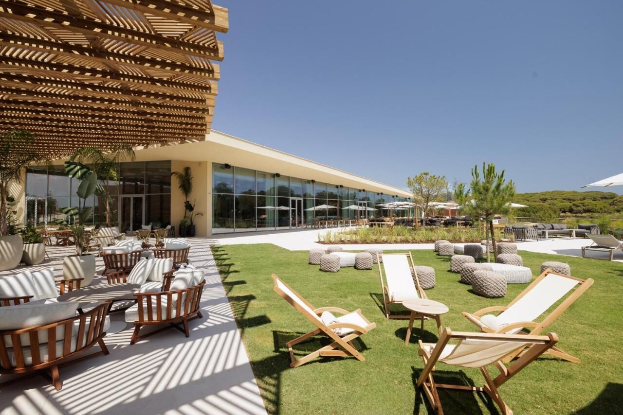 Un área de estar al aire libre con sillones de lounge y mesas, una pérgola de madera y un edificio con amplios ventanales.