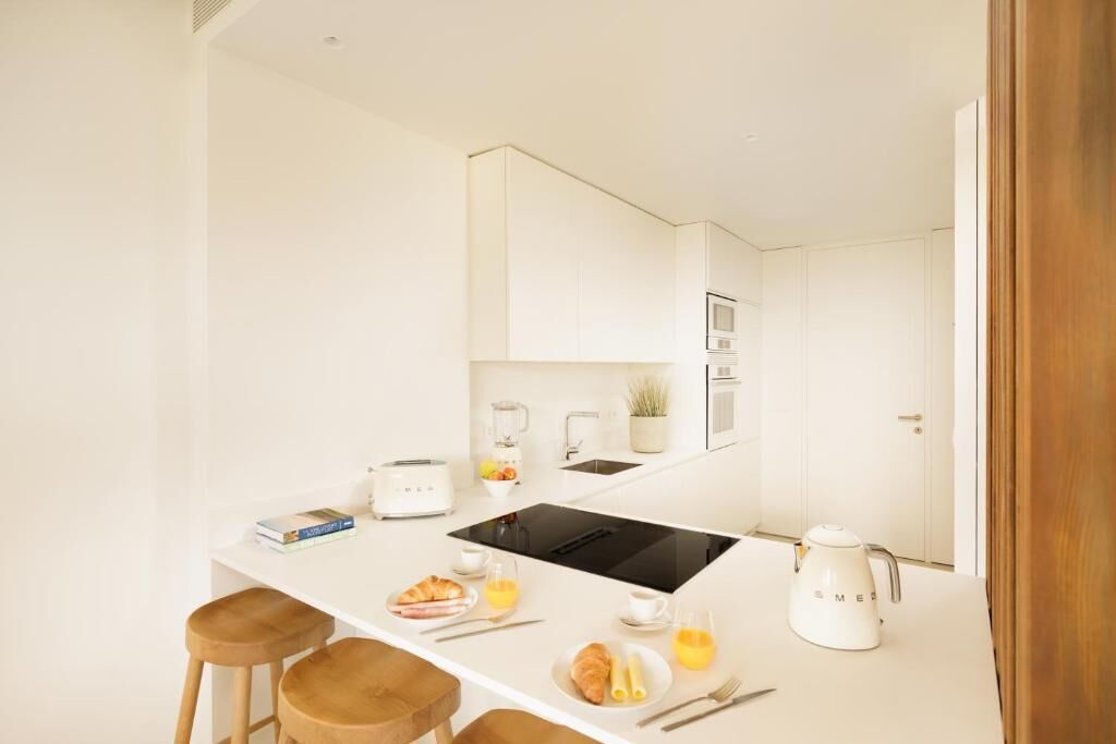 Una cocina moderna con armarios blancos, una cocina por inducción negra y un hervidor blanco. Hay taburetes de madera, una mesa pequeña con alimentos para el desayuno y una encimera blanca.