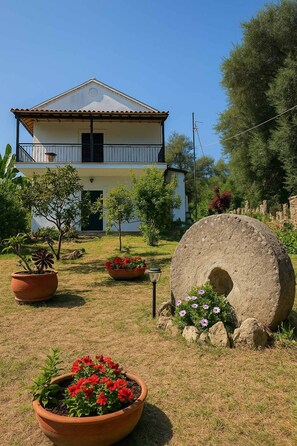 A two-story house with a balcony, surrounded by potted plants and a large stone structure.