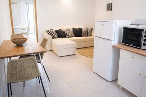 Eine kleine Küche mit Kühlschrank, Mikrowelle und kleinem Backofen. Ein Esstisch mit Stühlen und ein gewebter Korb sind vorhanden. Im Hintergrund befindet sich eine Couch mit gemusterten Kissen.