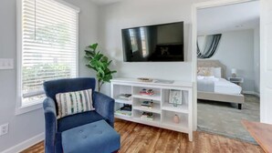 A room with a blue armchair, a white TV stand with books, a bed, and a large window with blinds. - The Tea Olive Cottage, Quaint | Pet-Friendly! (Savannah)