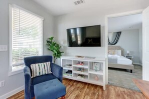 A modern bedroom with a large bed, a TV mounted on the wall, a blue armchair, and a white console table with books. - The Tea Olive Cottage, Quaint | Pet-Friendly! (Savannah)