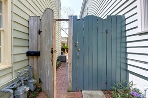 A gated backyard with a brick pathway leading to a utility area with pipes and an air conditioning unit. - The Tea Olive Cottage, Quaint | Pet-Friendly! (Savannah)