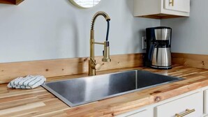 A kitchen with a stainless steel sink, a gold faucet, a coffee maker, and wooden cabinetry. - The Tea Olive Cottage, Quaint | Pet-Friendly! (Savannah)