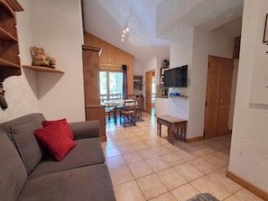 A living room with a sofa, red cushions, a television on the wall, wooden shelves, and a kitchen area in the background.