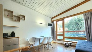 A room with a bed, a desk with a computer, a chair, a table with chairs, and a window with a view of trees. - 20m from ski slopes, 2nd floor, balcony, tv, 25m², Les Arcs (Les Arcs)