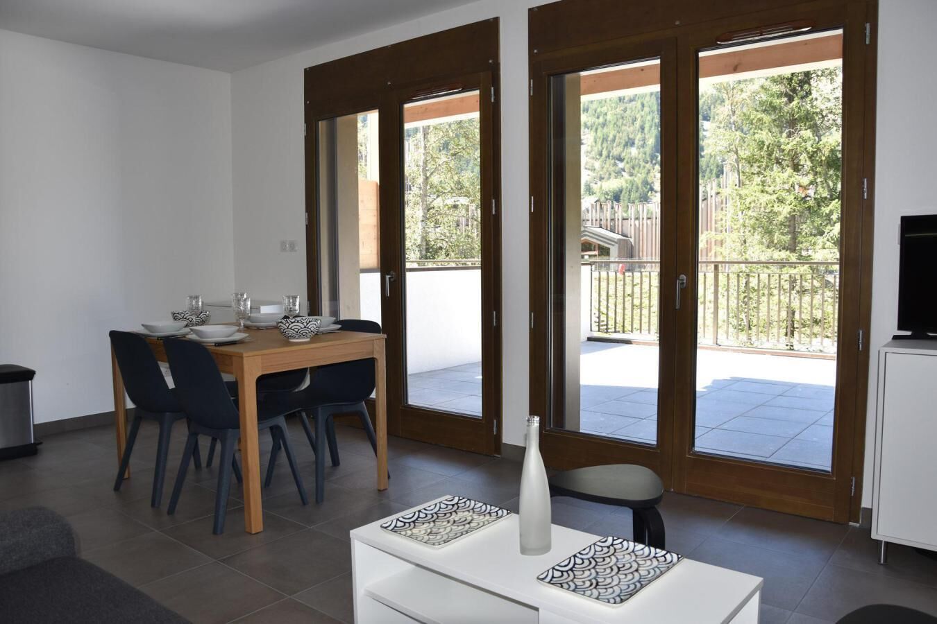 Zona pranzo con tavolo e sedie in legno, un armadietto bianco e un balcone affacciato su spazi verdi e un recinto.