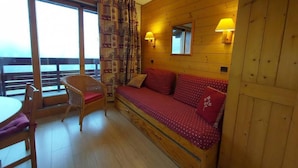 A cozy room with a wooden wall, a red sofa with patterned cushions, a wicker chair, a table, and a large window with curtains. - 3rd floor, view ski slopes, balcony, tv, ski locker, 13m², Valmorel (Valmorel)
