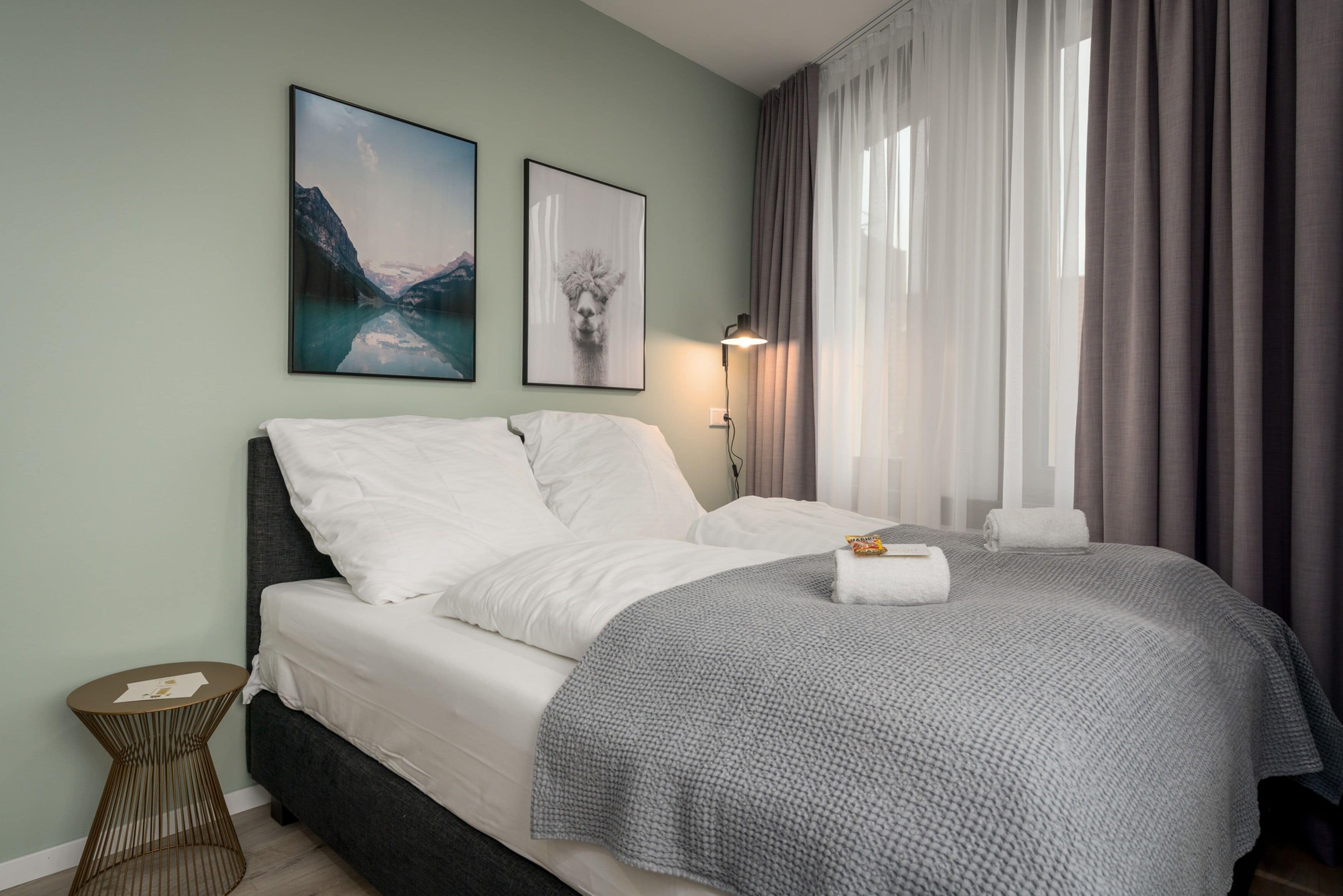 Un letto rifatto con lenzuola bianche, una coperta grigia e due asciugamani arrotolati. Due quadri appesi alla parete, un comodino e una lampada accanto al letto.