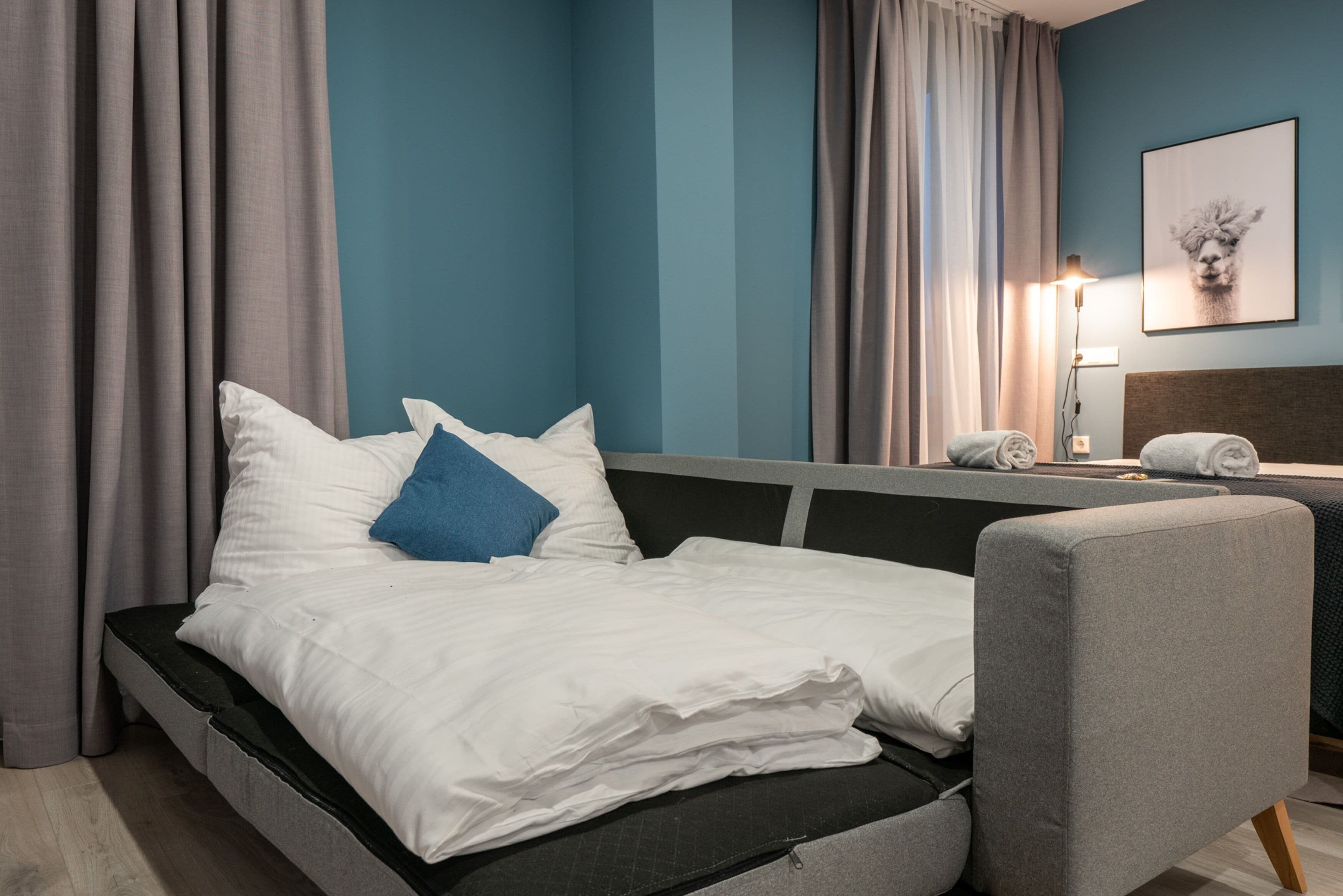 A hotel room with a bed, a chair, a nightstand, a lamp, and a wall-mounted picture.