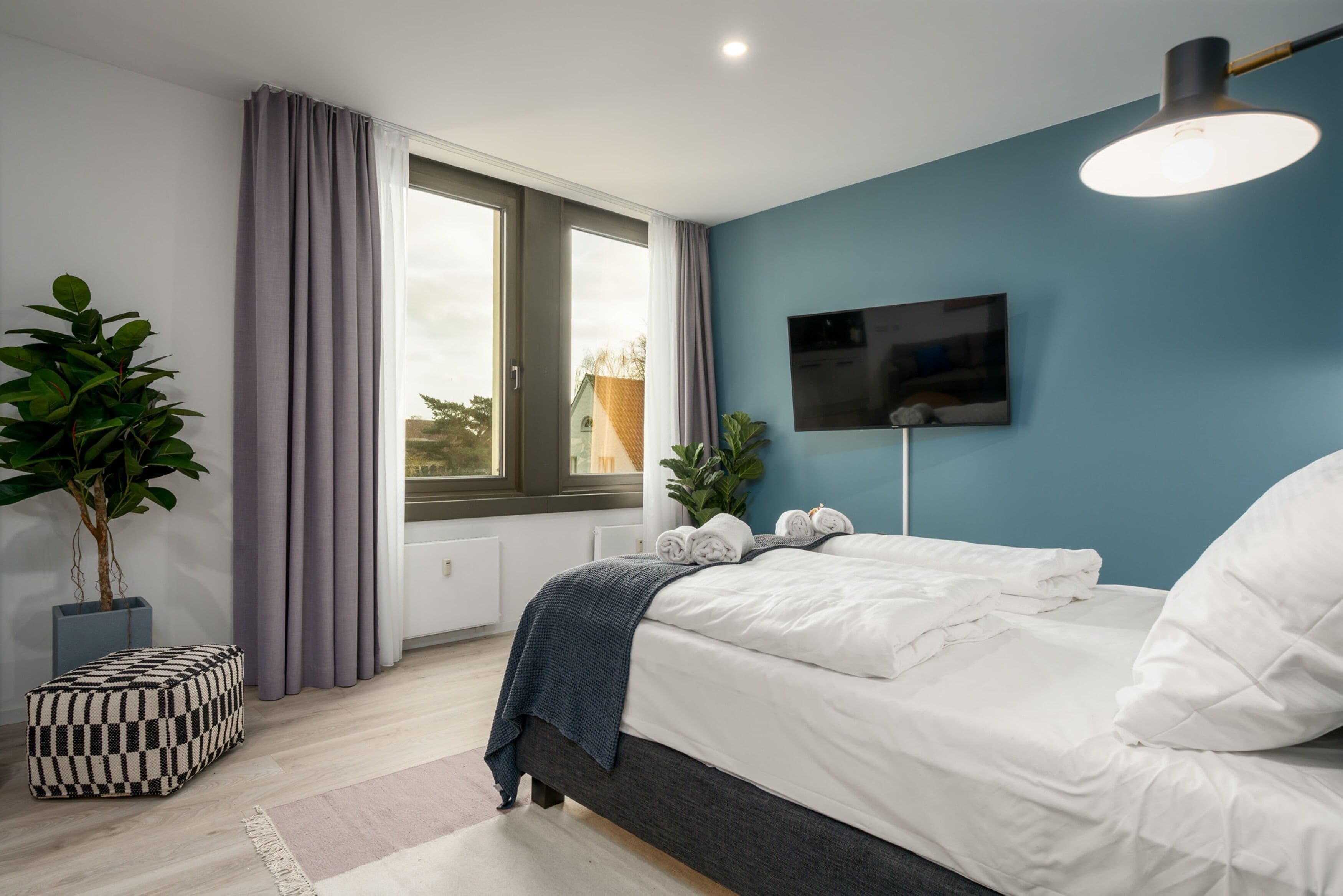 A modern bedroom with a large bed, a TV mounted on the wall, and a window with curtains.