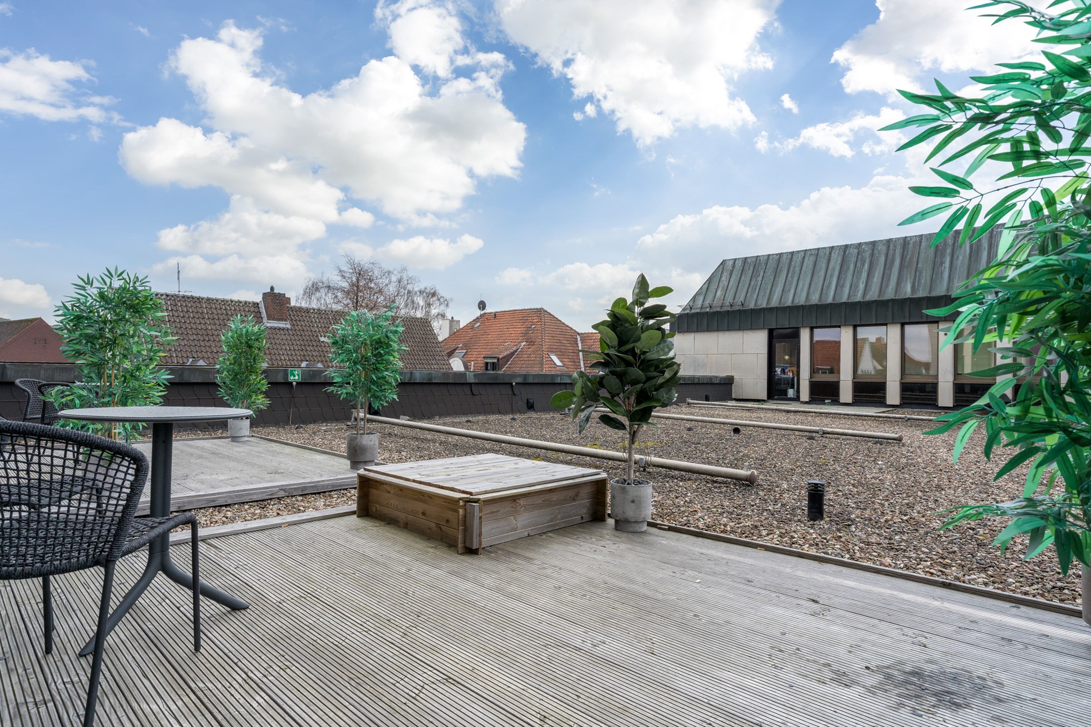 A rooftop patio with a table and chairs, surrounded by potted plants and a wooden deck.