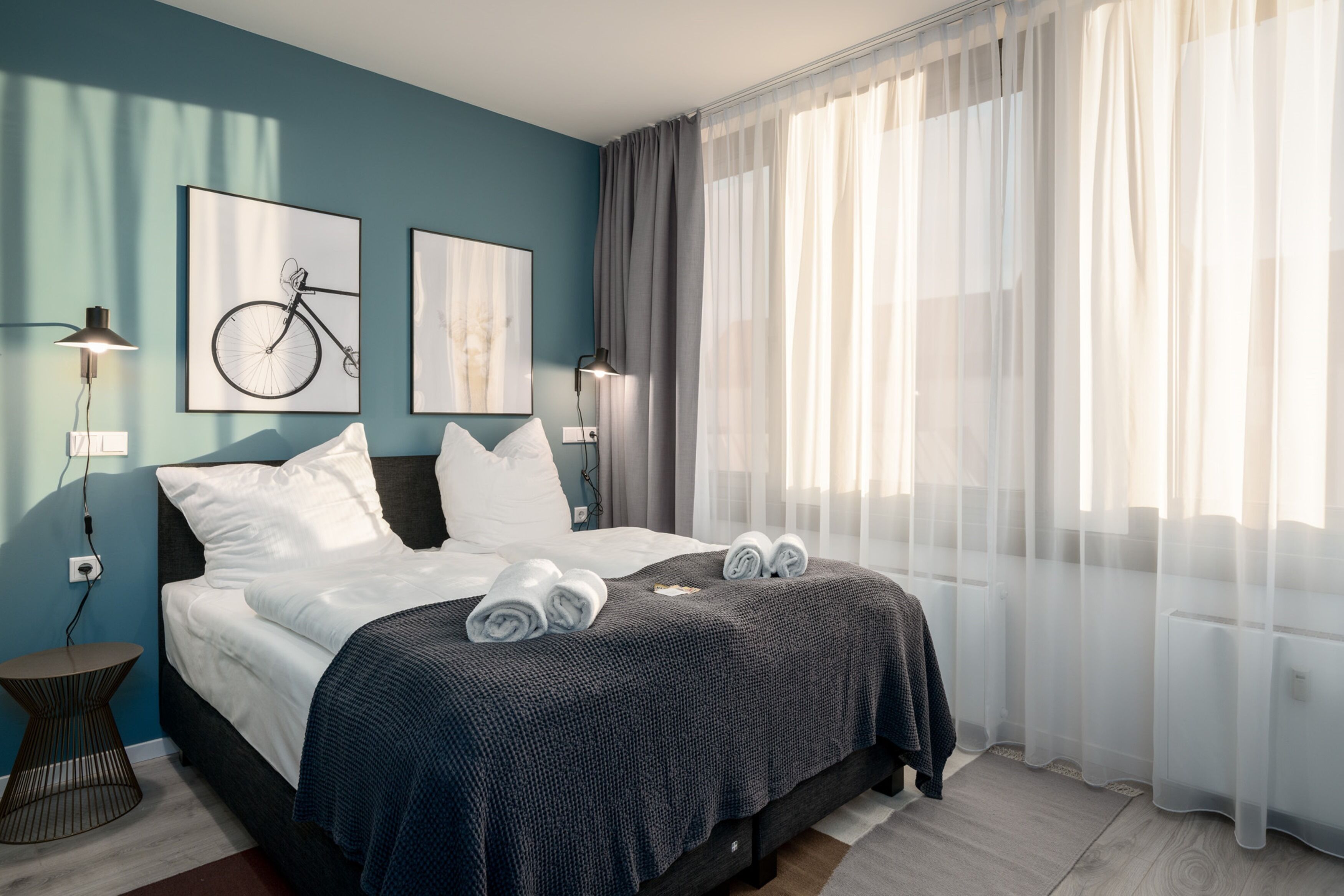 Une chambre d’hôtel moderne, équipée d’un lit, de deux lampes fixées au mur, d’une œuvre d’art représentant un vélo et d’une grande fenêtre avec des rideaux légers.