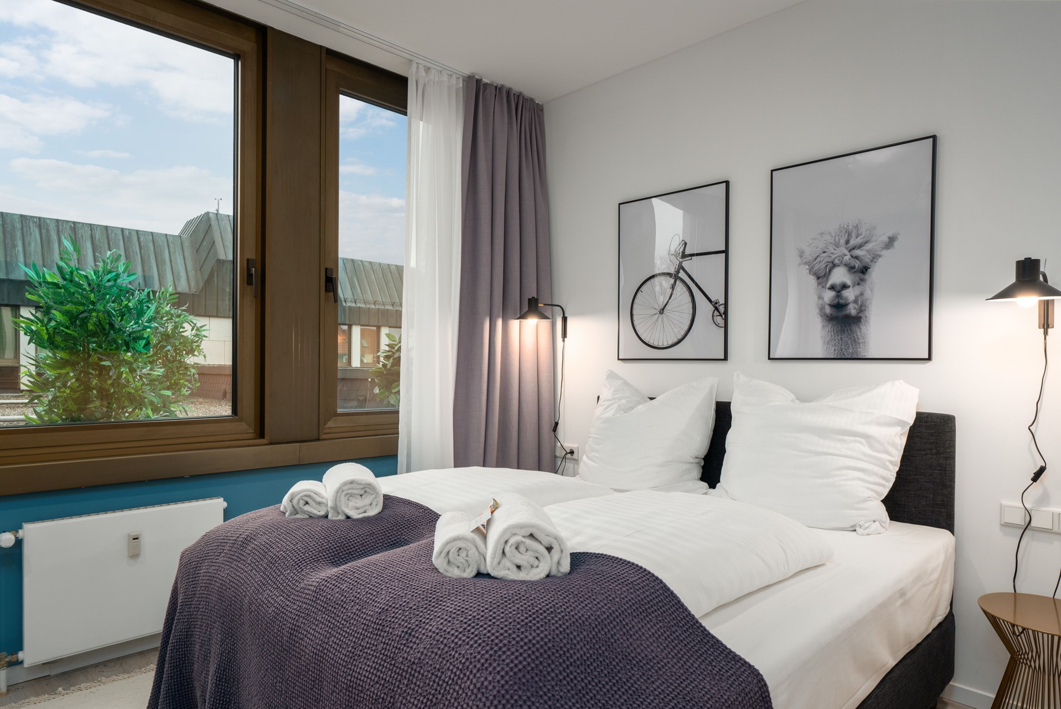 Une chambre d’hôtel avec une grande fenêtre, un lit recouvert de draps blancs, deux tableaux encadrés au mur et une lampe de chevet.