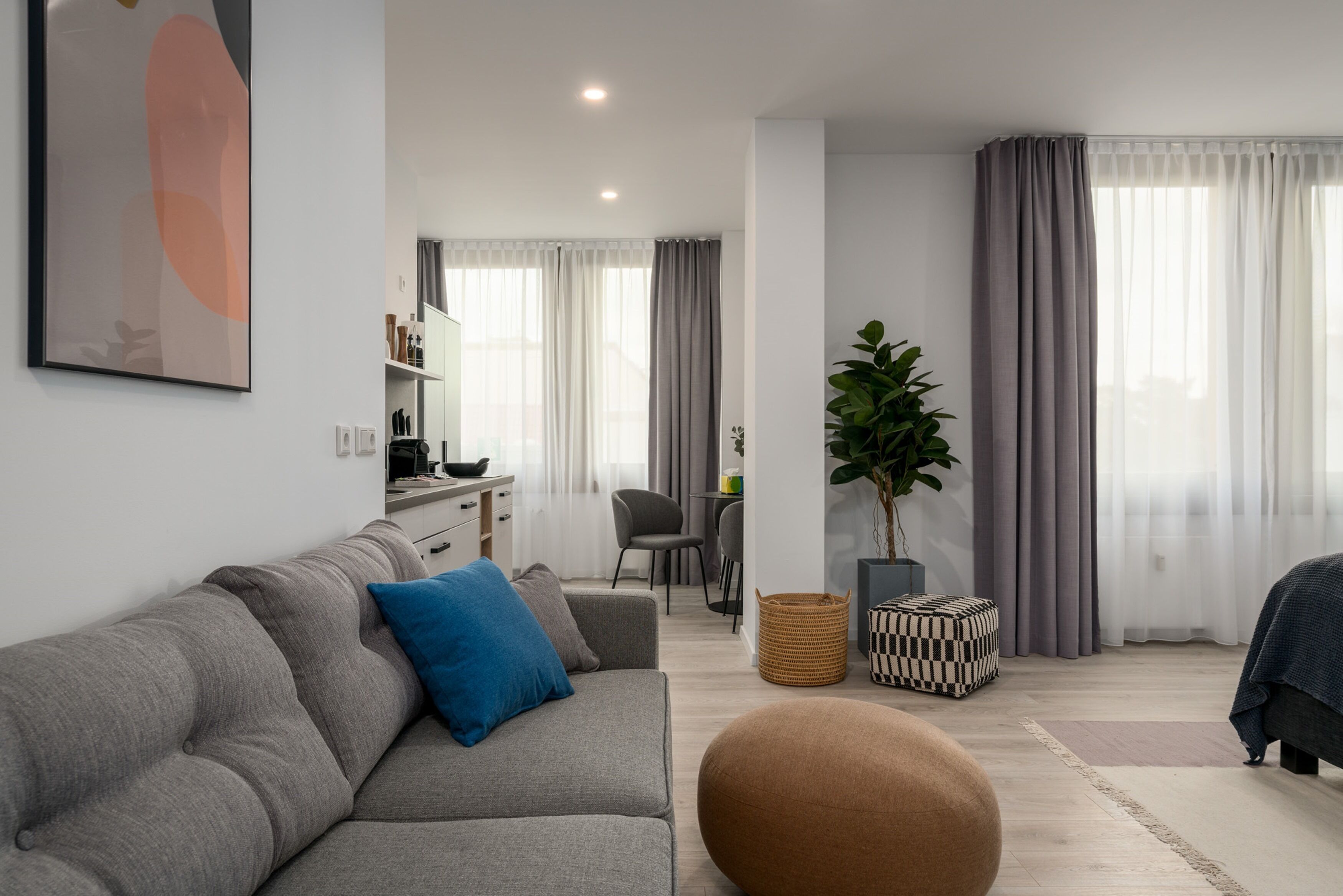 A modern living room with a grey sofa, a round ottoman, a potted plant, and a dining area in the background.