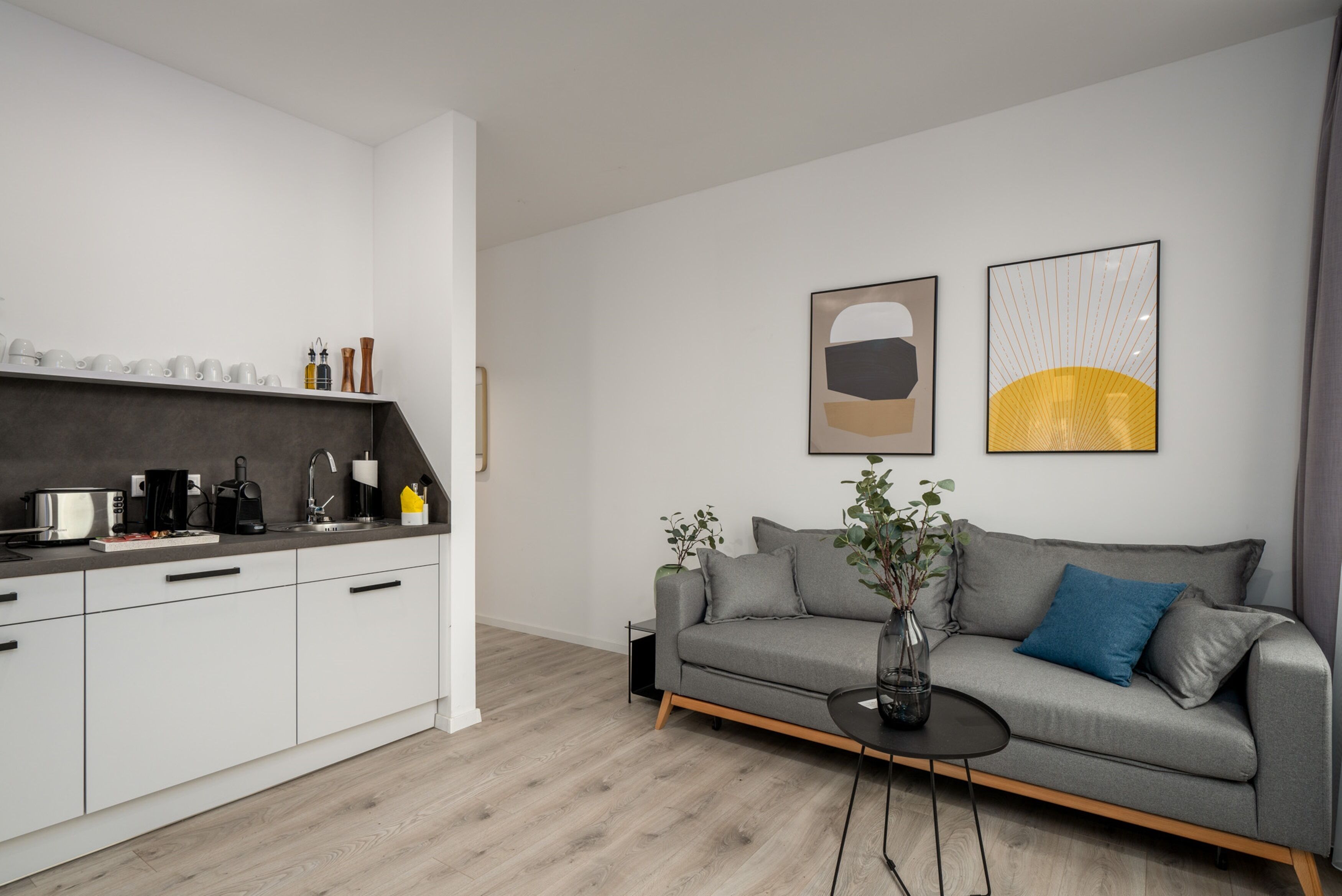 A modern living room with a grey sofa, a coffee table, and a kitchen area with white cabinets and a grey countertop.