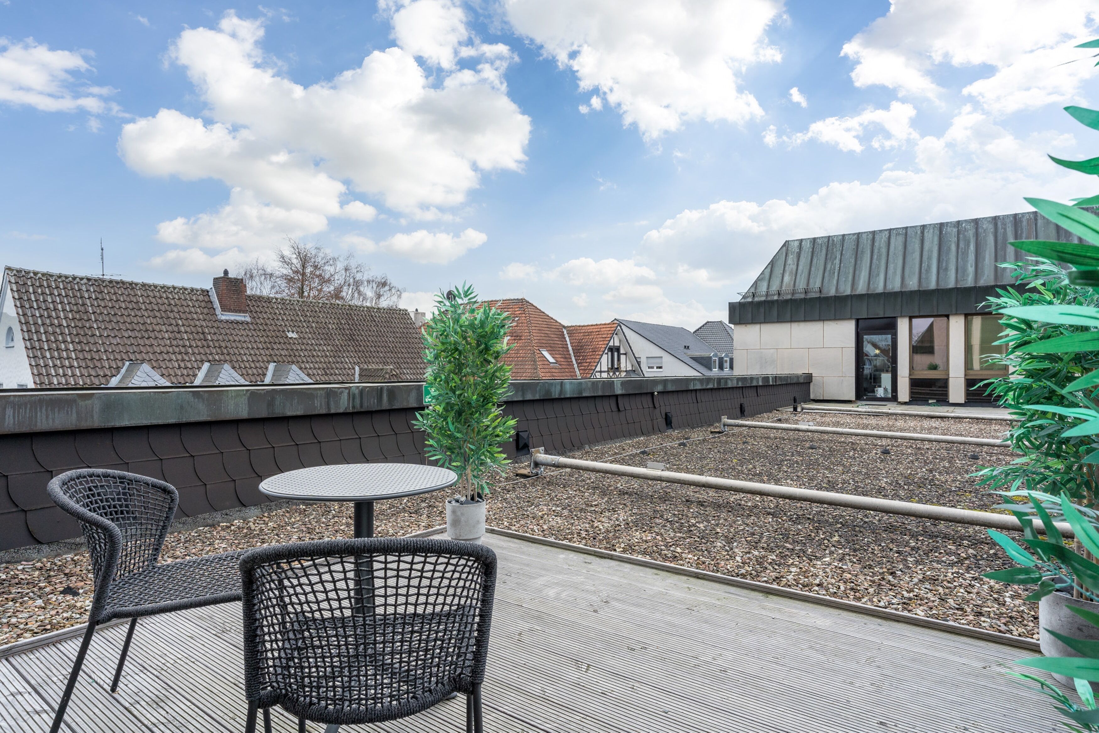 A rooftop terrace with a small table and chairs, a potted plant, and a view of buildings in the background.