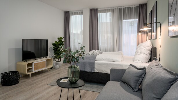 A modern living room with a flat-screen TV, a sofa, a coffee table, a plant, and a bed with curtains.