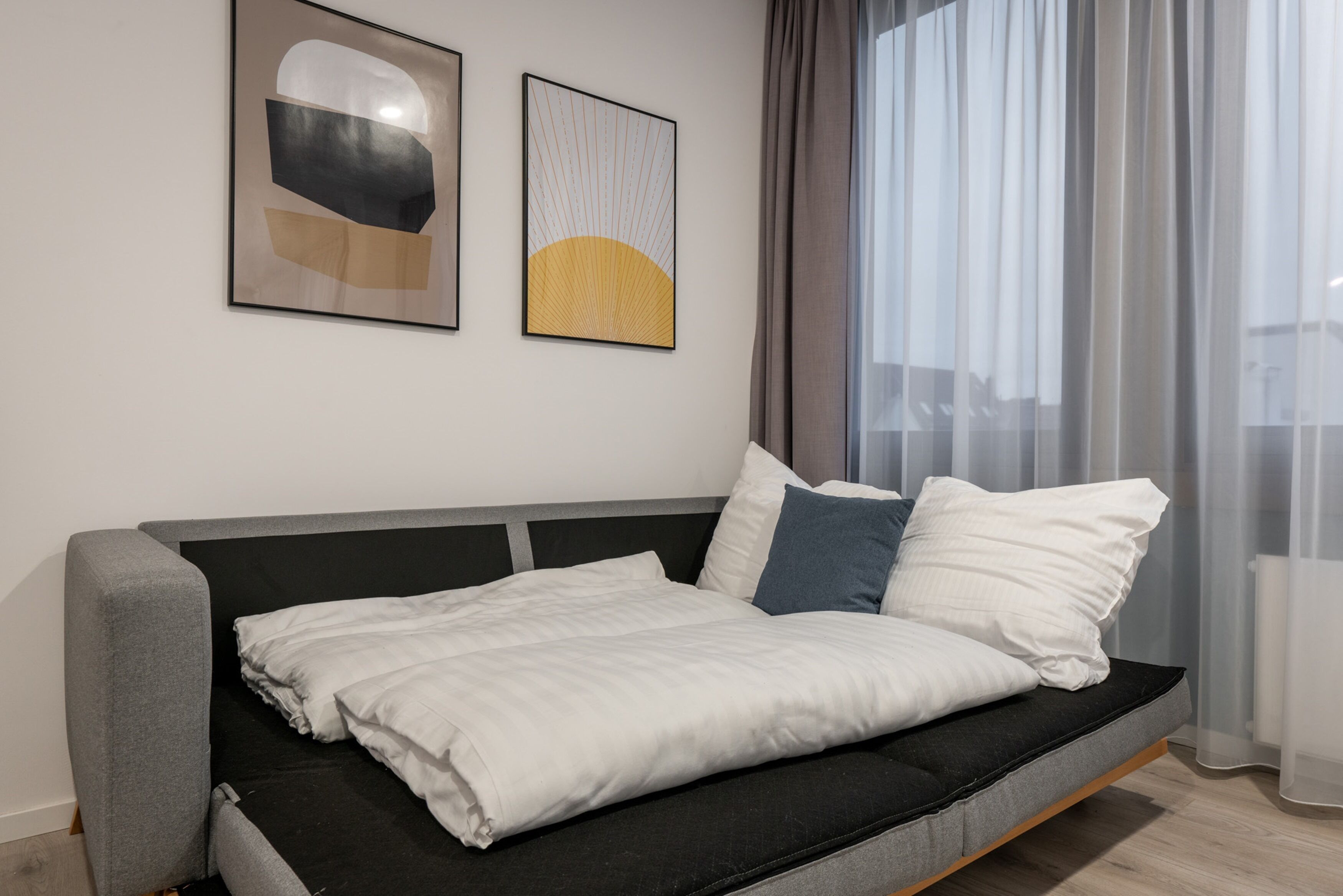A modern bedroom with a grey sofa bed, white and blue pillows, and two abstract wall art pieces.