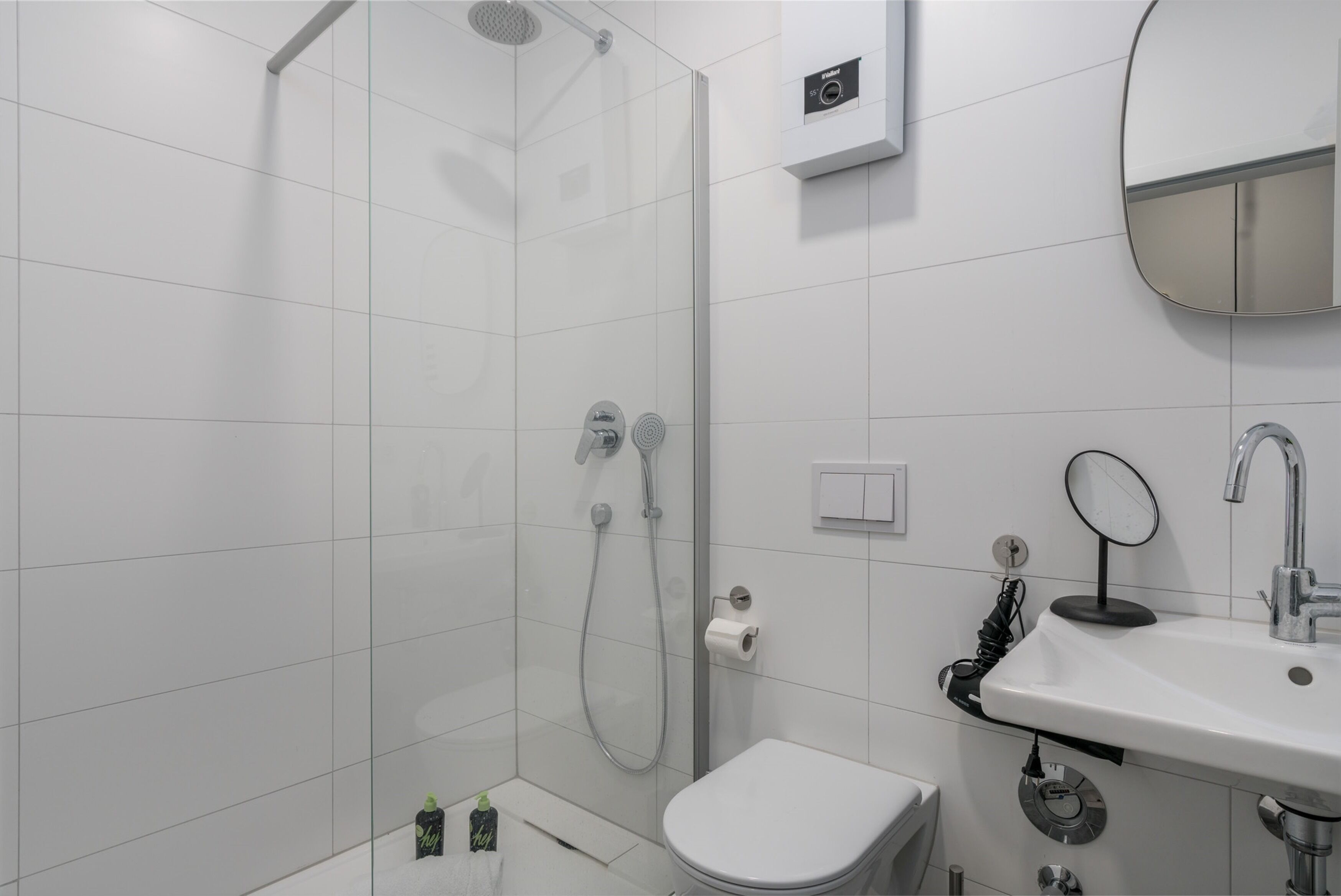 Un baño moderno con ducha envuelta en vidrio, inodoro blanco, lavabo con espejo e interruptor de luz en la pared.