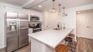 A modern kitchen with white cabinetry, stainless steel appliances, a large island with stools, and pendant lighting. - Covington Charm Suites (Covington)