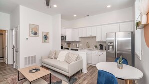 A modern kitchen with white cabinets, stainless steel appliances, and a dining area with a white table and blue chairs. - Covington Charm Suites (Covington)