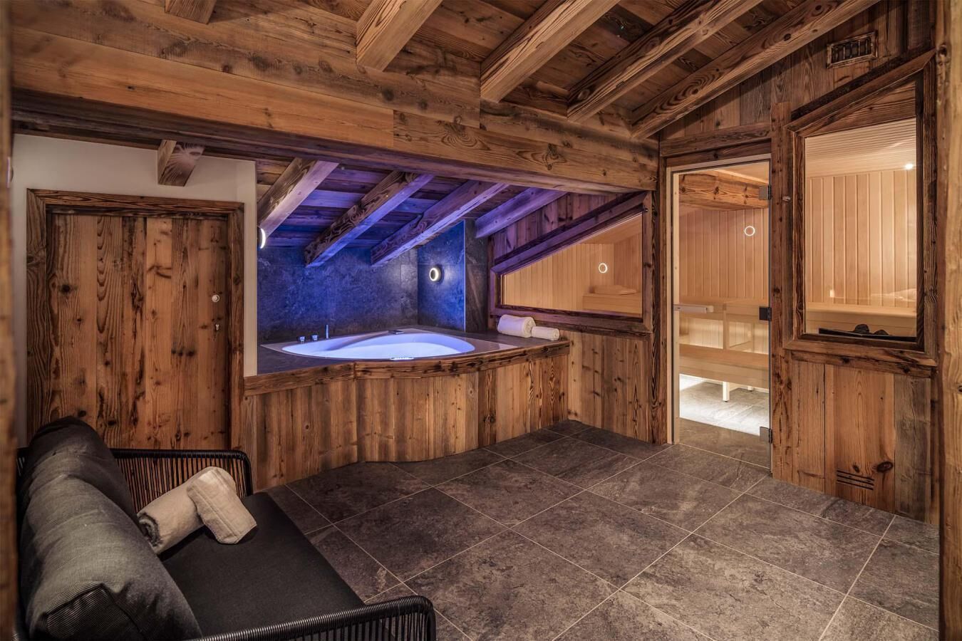 A wooden cabin with a stone-tiled floor, a wooden bench, and a large window overlooking a spa area.