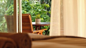 Quarto com cadeira de madeira, uma mesinha com planta em vaso e vista para o verde lá fora.
