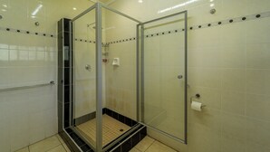 Cabina de ducha con paredes de vidrio, suelo de baldosas, un secador de manos y dispensador de papel higiénico en la pared.