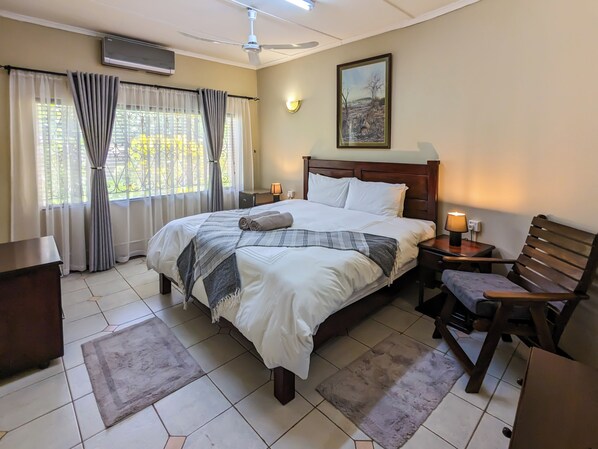 A hotel room with a bed, a desk, a chair, a nightstand, an air conditioner, a ceiling fan, a window with curtains, and a painting on the wall.
