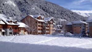 A snow-covered resort with multi-story buildings, a ski slope in the background, and a clear sky.