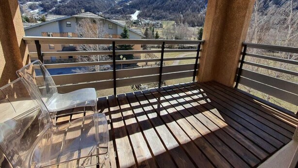A balcony with a wooden floor and metal railing, overlooking a mountainous landscape with buildings in the distance.
